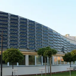 Elewacja budynku z panelami fotowoltaicznymi zamontowanymi na całej powierzchni ściany, widoczne okna i małe drzewka przed budynkiem oraz tabliczki z numerami.