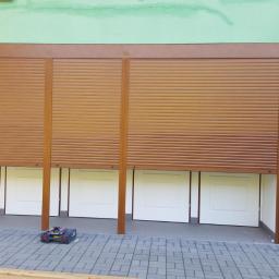 Technic Ml dystrybucja - Brązowe rolety zewnętrzne częściowo opuszczone na tle turkusowej ściany budynku, widoczny fragment chodnika z kostki brukowej i skrzynka z narzędziami.