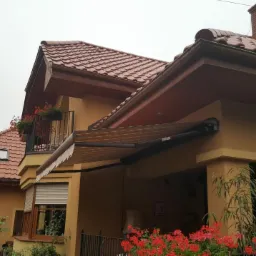 Zewnętrzna, brązowa markiza tarasowa zamontowana przy żółtym domu z czerwoną dachówką. Częściowo zwinięta roleta zewnętrzna na oknie. W tle balkon z kwiatami.