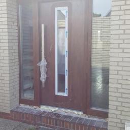 Technic Ml dystrybucja - Brązowe drzwi wejściowe z bocznymi przeszkleniami, jeszcze z folią ochronną na klamce i niebieskimi strzałkami na szybie, osadzone w elewacji z jasnej cegły.