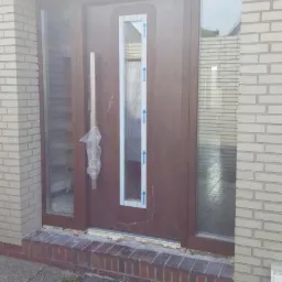 Brązowe drzwi wejściowe z bocznymi przeszkleniami, jeszcze z folią ochronną na klamce i niebieskimi strzałkami na szybie, osadzone w elewacji z jasnej cegły.