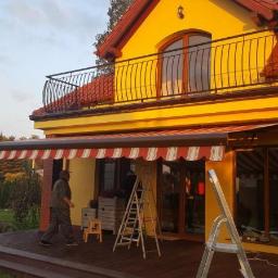 Technic Ml dystrybucja - Montaż markizy zewnętrznej w biało-czerwone paski przy żółtym domu z drewnianym tarasem, widoczne drabiny i pracownik w trakcie instalacji.