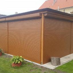 Technic Ml dystrybucja - Brązowe rolety zewnętrzne zamontowane na garażu w ogrodzie, widoczne donice z roślinami i fragment budynku w tle.