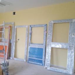 Technic Ml dystrybucja - Wnętrze pomieszczenia w trakcie remontu z żółtymi ścianami, gdzie widoczne są przygotowane do montażu ramy drzwiowe i okienne, niektóre jeszcze w folii ochronnej, oraz drabina i stół roboczy...