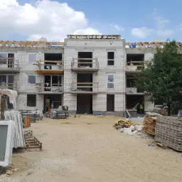 Wielorodzinny budynek w stanie surowym otwartym z widocznymi oknami PCV przygotowanymi do montażu, składowanymi na placu budowy.