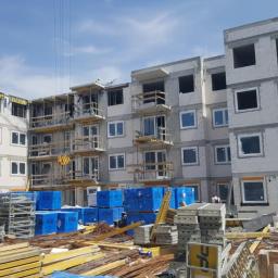 Technic Ml dystrybucja - Wielopiętrowy budynek w trakcie budowy z zamontowanymi oknami, widoczne rusztowania i dźwig budowlany, materiały budowlane składowane na placu budowy.