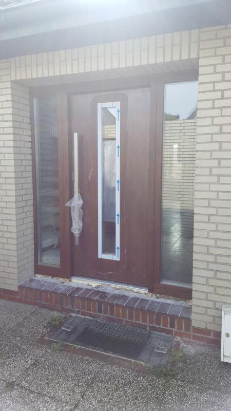 Brązowe drzwi wejściowe z bocznymi przeszkleniami, jeszcze z folią ochronną na klamce i niebieskimi strzałkami na szybie, osadzone w elewacji z jasnej cegły.