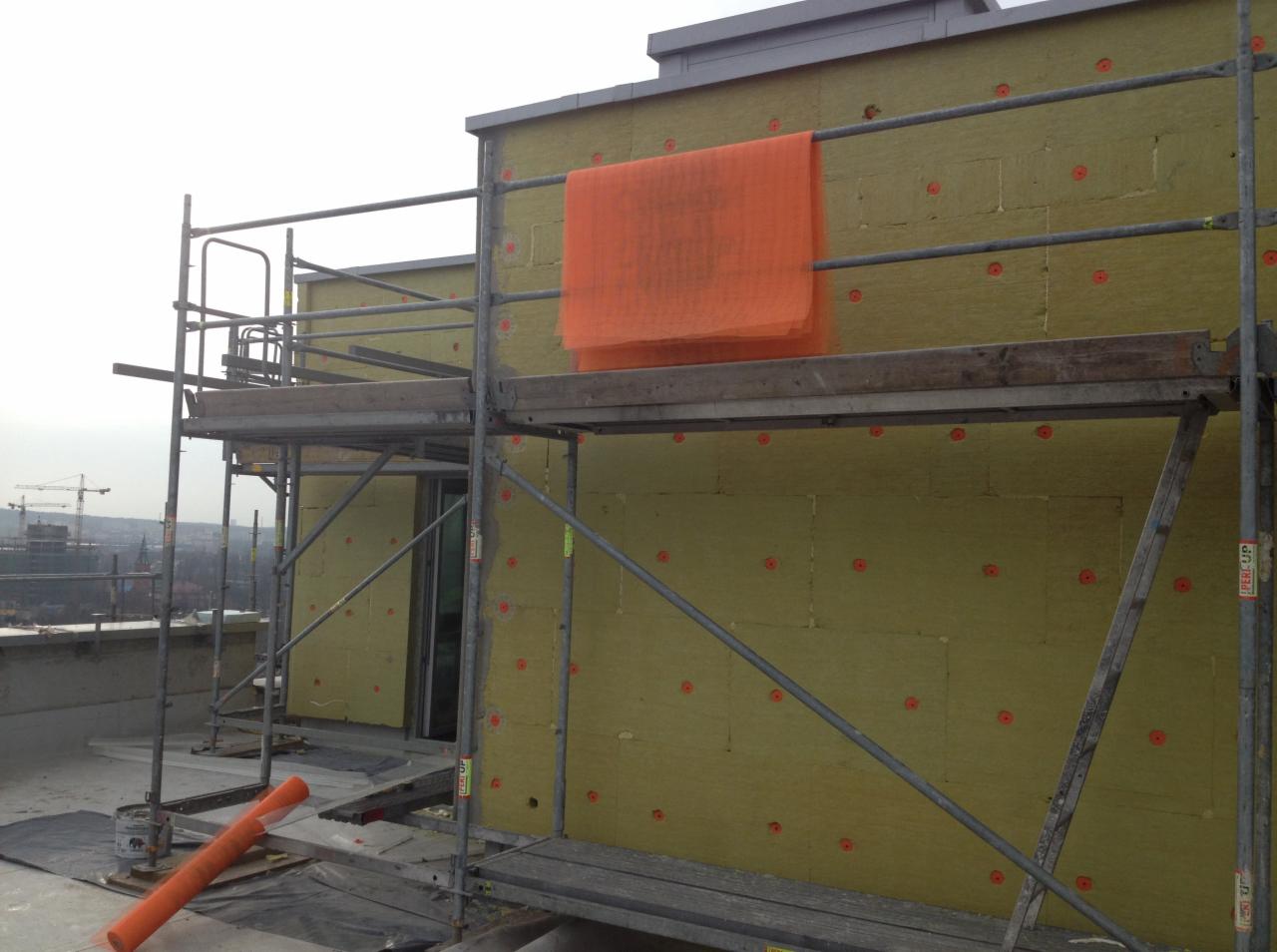 Ocieplanie budynku wełną mineralną, widoczne rusztowania i pomarańczowa siatka. Budynek w trakcie prac ociepleniowych z zewnątrz.
