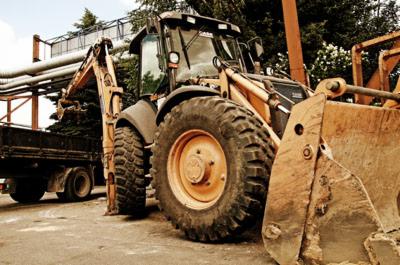 Żółto-czarna koparko-ładowarka z łyżką i przyczepą z rurami w tle, zaparkowana na placu budowy. Widoczne ślady użytkowania, oświetlenie dzienne.
