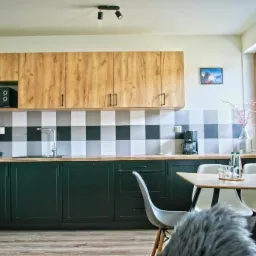 Nowoczesna kuchnia z ciemnozielonymi szafkami dolnymi i drewnianymi górnymi, wzorzyste płytki na ścianie, stół z krzesłami, dekoracje i oświetlenie punktowe.