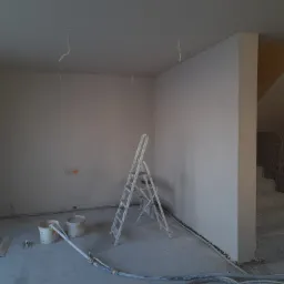 Pokój w trakcie prac wykończeniowych: białe ściany, schody z surowego betonu z widoczną fakturą, drabina malarska, wiaderka z zaprawą i przewody do agregatu tynkarskiego leżące na podłodze.