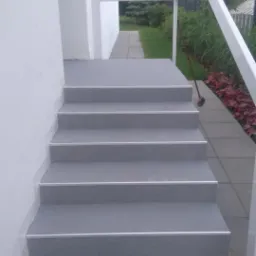 Zewnętrzne schody pokryte szarą żywicą z białymi listwami krawędziowymi, prowadzące do wejścia z białą ramą i widokiem na zielony ogród.