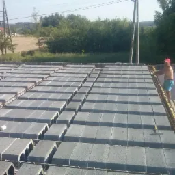 Wypełniony strop Teriva z betonowych pustaków, widoczny pracownik w czerwonej czapce przy wznoszonej ścianie, krajobraz wiejski w tle.