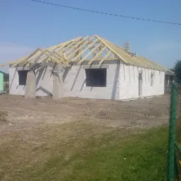Budowa domu jednorodzinnego: wzniesione ściany z betonu komórkowego i drewniana konstrukcja dachu, widok z zewnątrz na plac budowy w słoneczny dzień.
