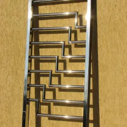 Konstrukcja ze stali nierdzewnej z geometrycznym wzorem, prawdopodobnie element balustrady lub poręczy, ustawiona na tle teksturowanej ściany w ciepłym odcieniu beżu.