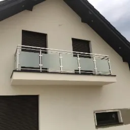Nowoczesny balkon z przeszkloną balustradą i nierdzewnymi elementami mocującymi, widok z dołu na tle białej elewacji budynku z czarną stolarką okienną i dachem.