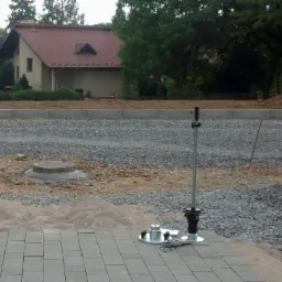 Badanie gruntu w toku: ubijak dynamiczny na kostce brukowej, grunt przygotowany pod budowę domu z czerwonym dachem w tle. Prace geologiczne w terenie.