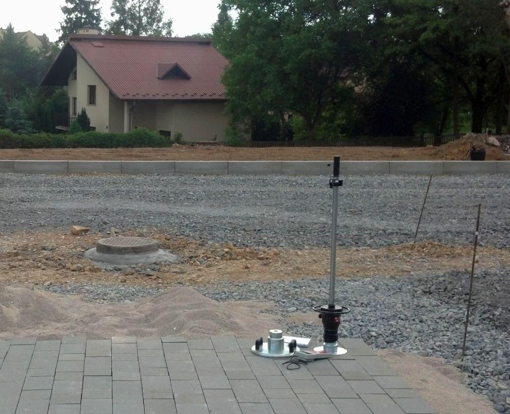 Badanie gruntu w toku: ubijak dynamiczny na kostce brukowej, grunt przygotowany pod budowę domu z czerwonym dachem w tle. Prace geologiczne w terenie.