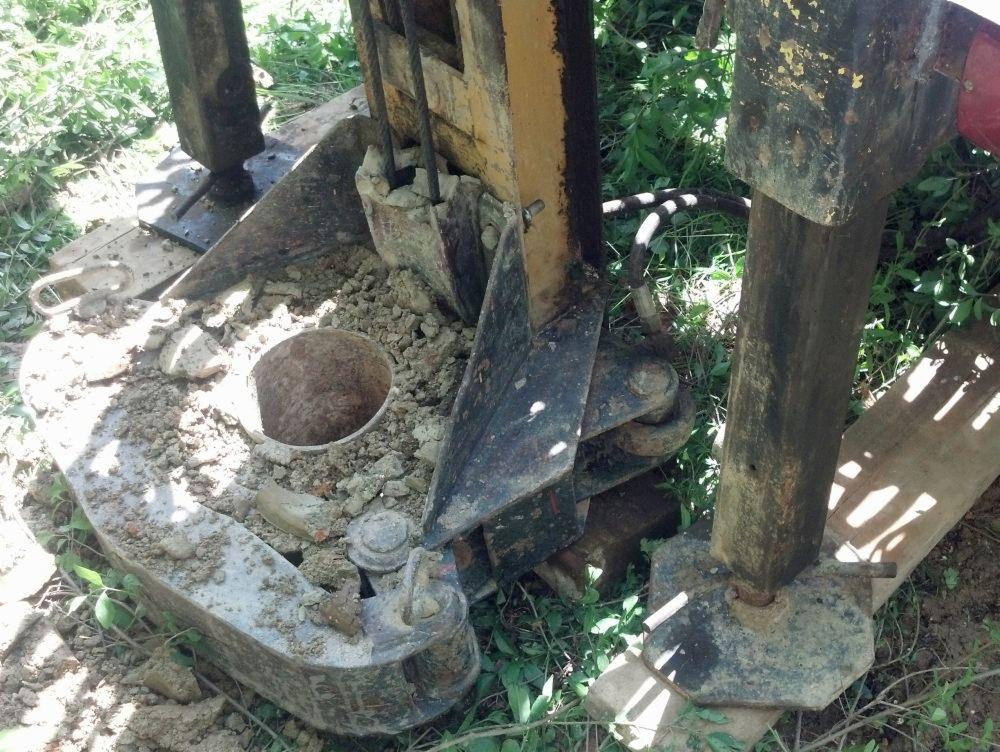 Wiertnica geologiczna w trakcie pracy, widoczny otwór w ziemi i fragment urządzenia w otoczeniu zieleni. Sprzęt z elementami rdzy.