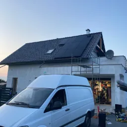 Montaż paneli fotowoltaicznych na dachu domu jednorodzinnego, widoczne rusztowanie i biały samochód dostawczy z narzędziami.