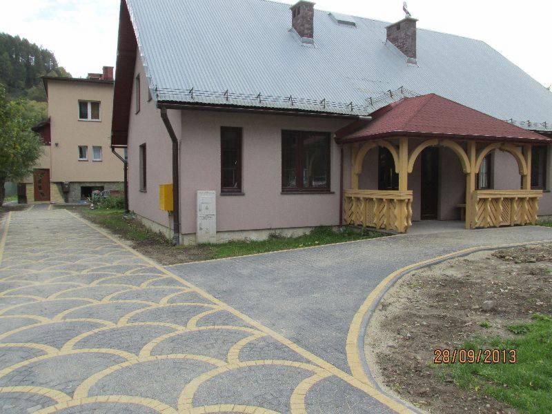 Szara kostka brukowa z żółtymi akcentami prowadzi do budynku z drewnianą werandą i szarym dachem. Widoczny fragment elewacji budynku obok.