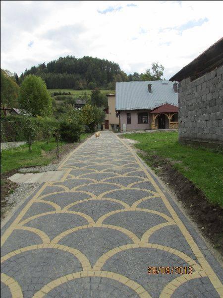 Ścieżka z kostki brukowej w odcieniach szarości i beżu, ułożona we wzór wachlarzy, prowadząca w stronę domu na tle zielonego wzgórza.