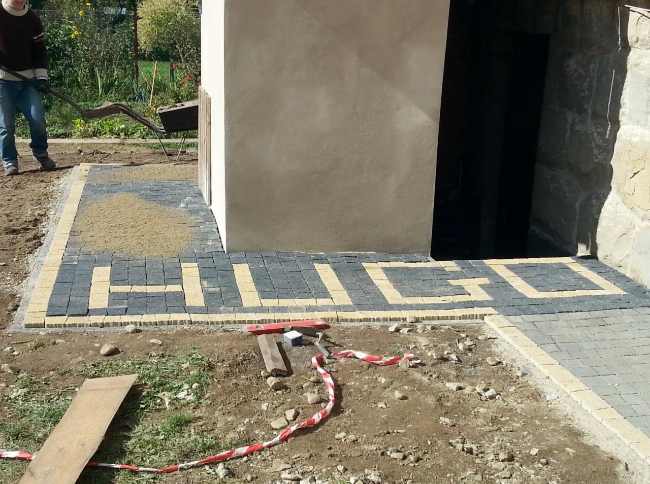 Układanie kostki brukowej z napisem 'HUGO' w żółto-szarej kolorystyce, widoczne narzędzia i fragment osoby z taczką. Prace brukarskie w toku.