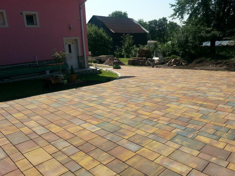 Kolorowa kostka brukowa ułożona na podjeździe przed różowym domem z białymi oknami i drzwiami. W tle widoczny budynek gospodarczy i drzewa.