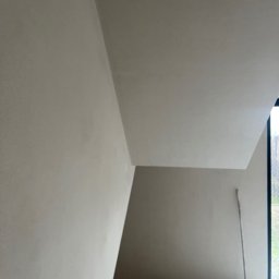 MBM Warszawa sp. z o.o. - Wnętrze w trakcie prac wykończeniowych, ściany i sufit pokryte świeżo nałożoną, jasną warstwą, widoczna szczotka oparta o ścianę przy oknie.