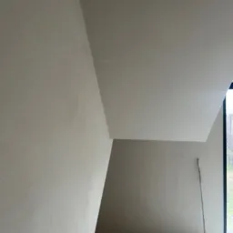 Wnętrze w trakcie prac wykończeniowych, ściany i sufit pokryte świeżo nałożoną, jasną warstwą, widoczna szczotka oparta o ścianę przy oknie.