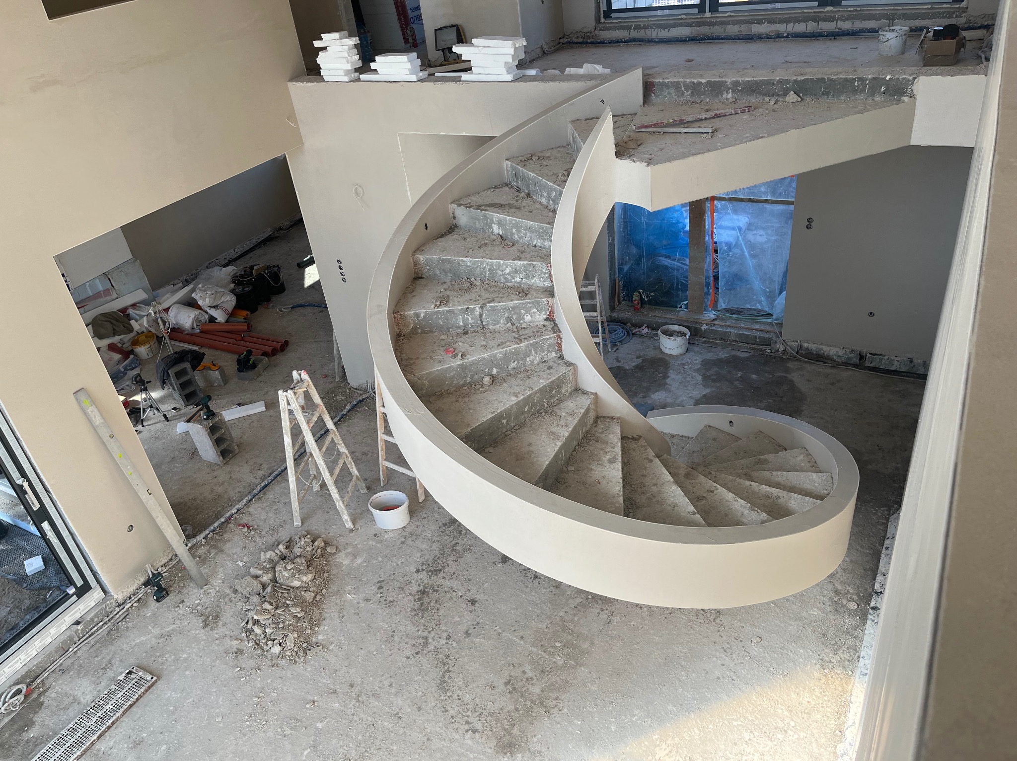 Wnętrze domu w trakcie prac wykończeniowych: spiralne schody z surowego betonu z białymi balustradami, świeżo otynkowane ściany w odcieniach beżu, drabina i narzędzia budowlane na podłodze.