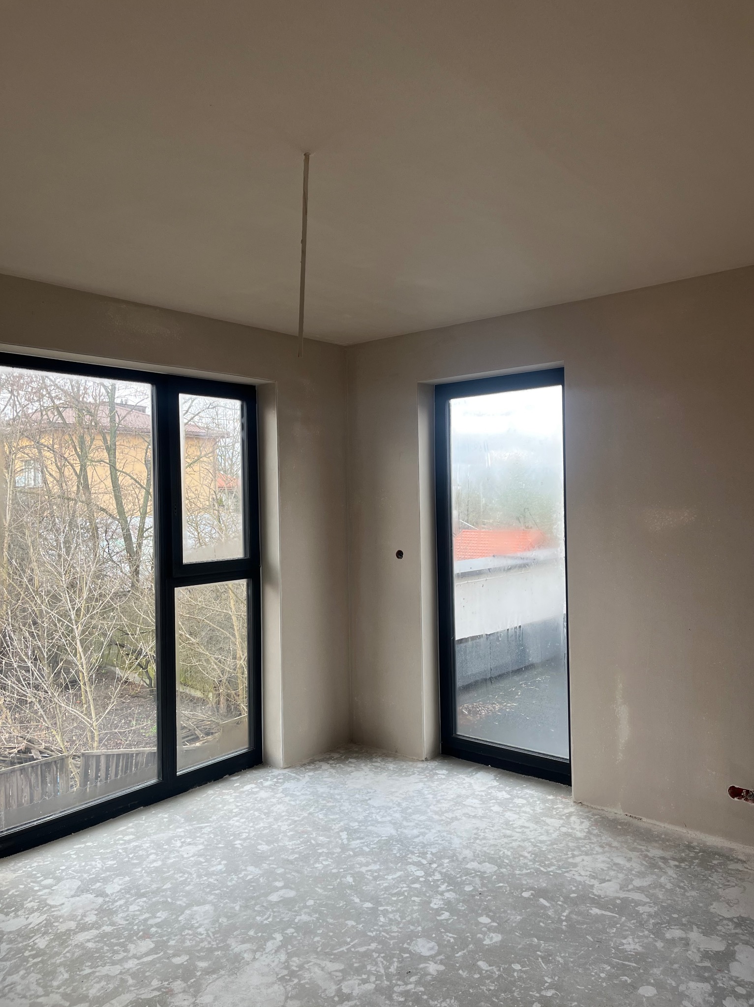 Pokój w stanie deweloperskim po nałożeniu gładzi, widoczne dwa okna w czarnych ramach i surowy beton na podłodze.