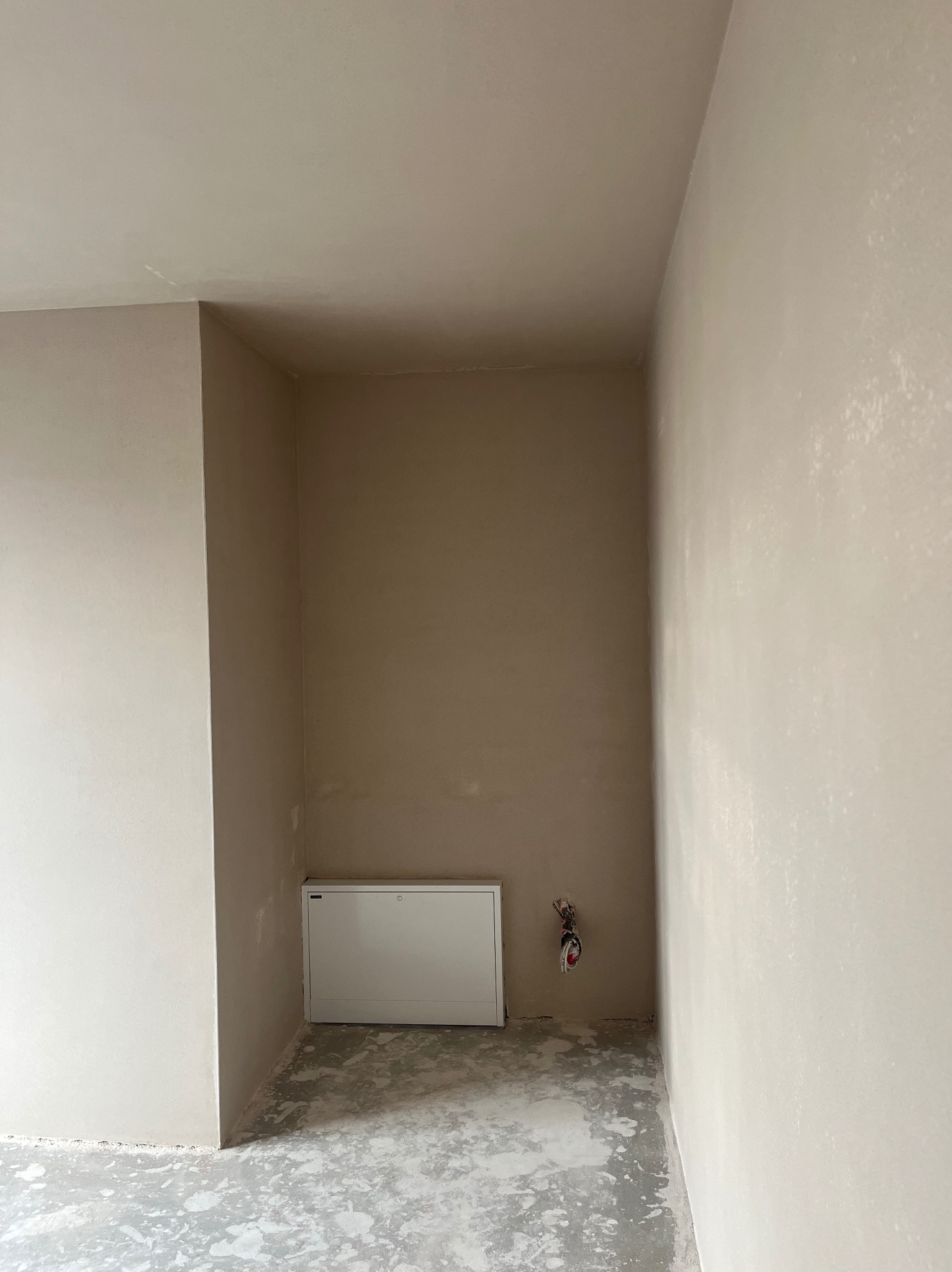 Wnętrze w trakcie prac wykończeniowych: ściany pokryte świeżą, jasnobeżową gładzią, widoczna biała skrzynka rewizyjna i wystające przewody, surowa wylewka na podłodze.