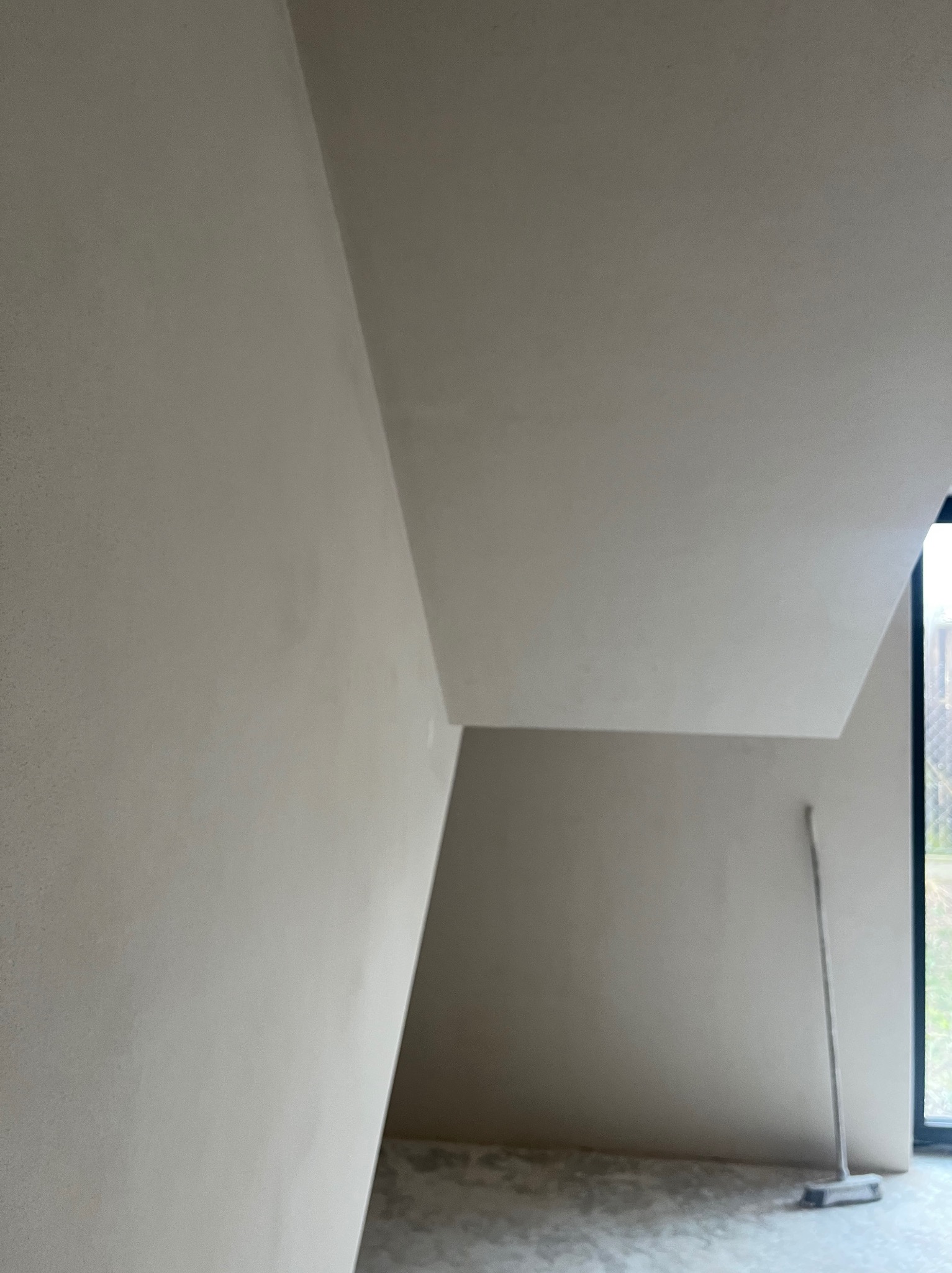 Wnętrze w trakcie prac wykończeniowych, ściany i sufit pokryte świeżo nałożoną, jasną warstwą, widoczna szczotka oparta o ścianę przy oknie.