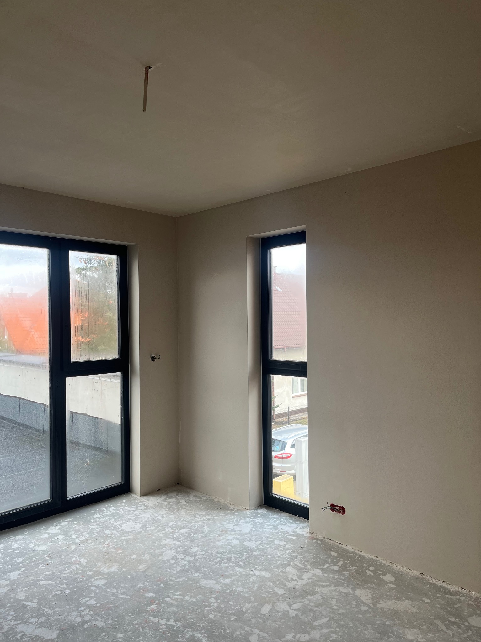 Wnętrze pokoju w stanie deweloperskim z oknami w czarnych ramach, ściany pokryte świeżo nałożoną warstwą gładzi, podłoga z resztkami zaprawy, widoczne wystające przewody elektryczne.