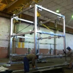 Spawanie metalowej konstrukcji szkieletowej hali w hali produkcyjnej pod suwnicą. Dwóch spawaczy przy pracy, jeden szlifuje element, drugi spawa.