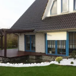 Pergola z drewnianym tarasem przylegająca do domu z cegły, obok oczko wodne z białymi kamieniami i roślinami. Dom z brązowym dachem.