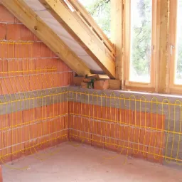 Żółte kable ogrzewania podłogowego ułożone na ceglanej ścianie w Warszawie, widoczne drewniane elementy konstrukcji dachu i okno. Etap instalacji przed wylaniem posadzki.