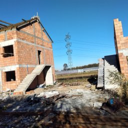 Bud-Dom - Wznoszenie dwóch budynków w stanie surowym otwartym z czerwonej cegły, z widocznymi betonowymi schodami zewnętrznymi, na tle błękitnego nieba i słupa wysokiego napięcia.