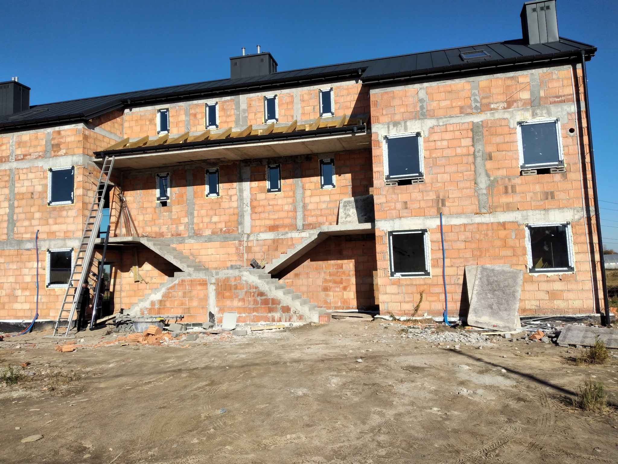 Budynek w stanie surowym otwartym z widocznymi schodami zewnętrznymi, częściowo wstawionymi oknami i metalową drabiną opartą o ścianę.