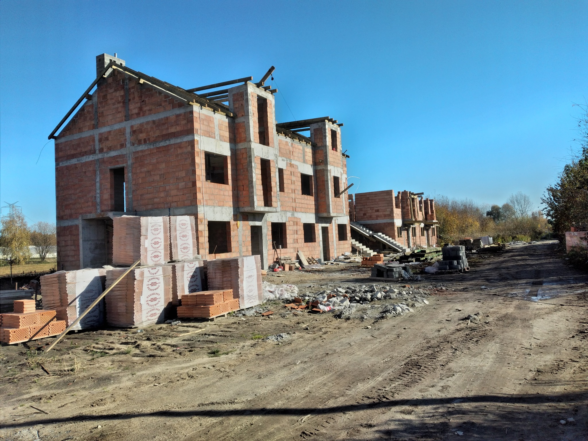 Wczesny etap budowy szeregu domów z czerwonej cegły, widoczne palety z materiałem budowlanym i wykopana droga dojazdowa.