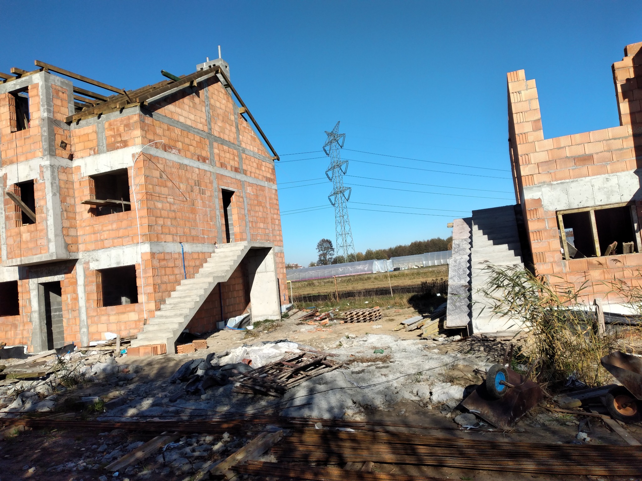 Wznoszenie dwóch budynków w stanie surowym otwartym z czerwonej cegły, z widocznymi betonowymi schodami zewnętrznymi, na tle błękitnego nieba i słupa wysokiego napięcia.