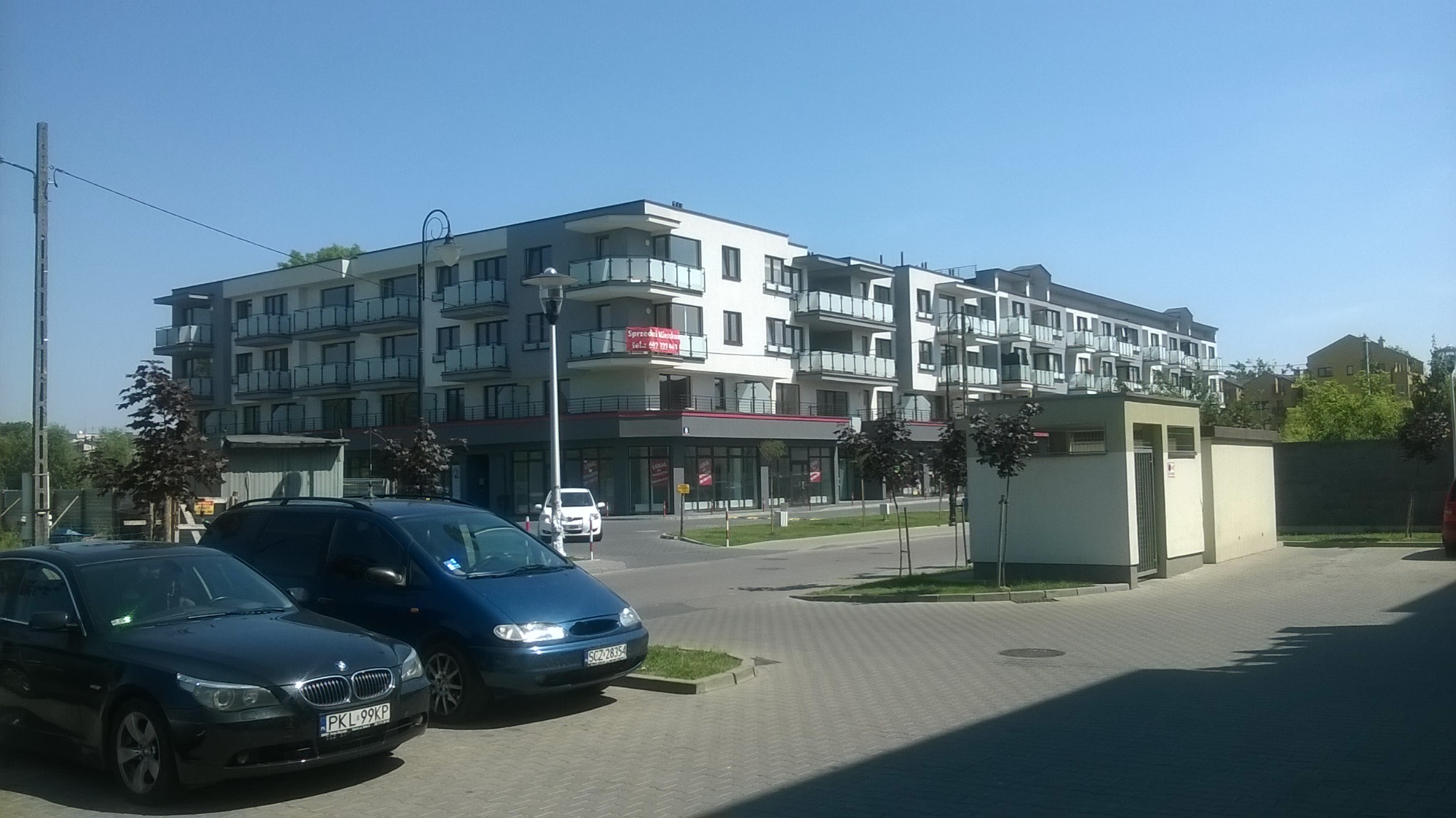Nowoczesny, wielopiętrowy budynek mieszkalny z balkonami i lokalami usługowymi na parterze, widoczny z poziomu ulicy, z zaparkowanymi samochodami na pierwszym planie i banerem reklamowym 'Sprzedaż...
