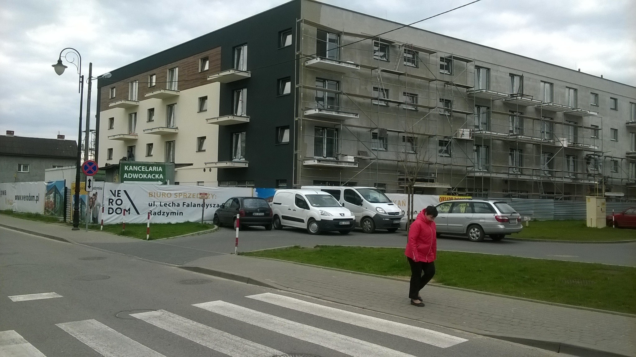 Wielopiętrowy budynek w trakcie wykończenia elewacji, widoczne rusztowania, zaparkowane samochody i przechodząca kobieta w czerwonej kurtce na pierwszym planie.