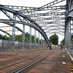 Stalowa konstrukcja mostu w trakcie remontu. Widoczna siatka zbrojeniowa na jezdni i pracownicy w odblaskowych kamizelkach. Charakterystyczne, ażurowe wzmocnienia konstrukcji.