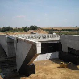 Betonowy most w budowie z widocznym zbrojeniem i izolacją. Mała koparka stoi na ziemi. W tle pola i drzewa pod błękitnym niebem.