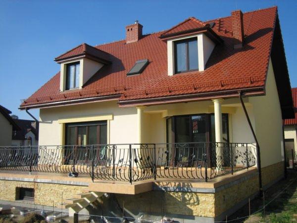 Dwupiętrowy dom z czerwonym dachem, balkonem z kutą balustradą i elewacją w odcieniach beżu w słoneczny dzień. Widoczne okna dachowe i komin z cegły.