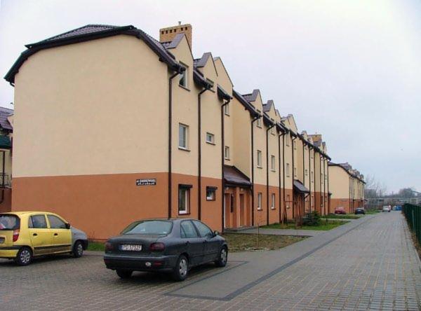 Rząd szeregowych domów z dwukolorową elewacją (beżowo-brązową) i brązowymi dachami w pochmurny dzień. Na pierwszym planie zaparkowane auta.