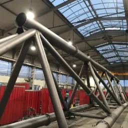 Wielkogabarytowa konstrukcja stalowa kratowa z rur, spawana w hali produkcyjnej, widok z dołu na sufit z oknami.