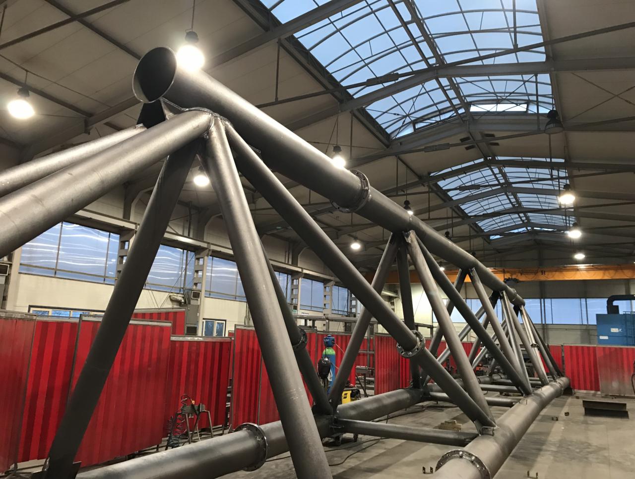 Wielkogabarytowa konstrukcja stalowa kratowa z rur, spawana w hali produkcyjnej, widok z dołu na sufit z oknami.